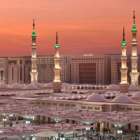 Intercontinental Dar Al Iman Madinah By Ihg Hotel Medina Eksteriør billede The photo depicts a scenic view of a mosque complex, likely during sunset, with several minarets illuminated and showcasing green lights. The architecture features domed roofs, and the setting includes a backdrop of buildings, all bathed in a warm, o