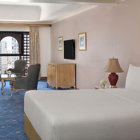 Intercontinental Dar Al Iman Madinah By Ihg Hotel Medina Eksteriør billede The photo shows a well-furnished hotel room. It features a large bed with a white duvet, two bedside lamps, and decorative paintings on the walls. There is a comfortable seating area with two chairs and a small table. A wooden dresser is positioned a
