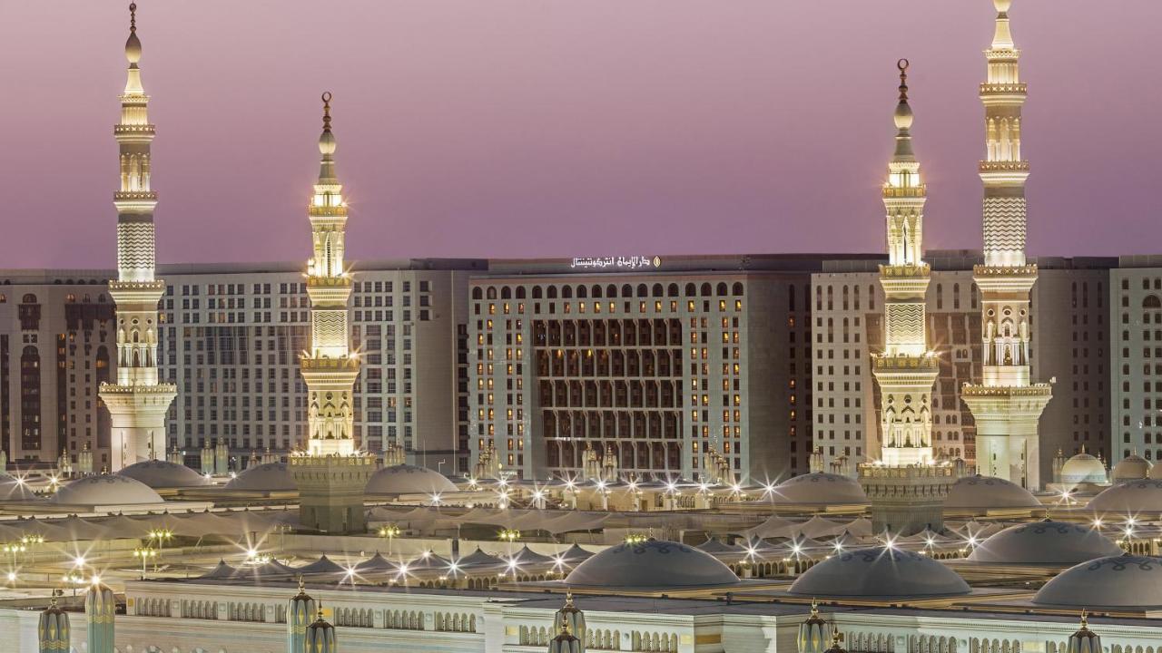 Intercontinental Dar Al Iman Madinah By Ihg Hotel Medina Eksteriør billede The photo depicts an architectural scene, likely from a mosque complex, featuring prominent minarets that are illuminated. The setting appears to be during twilight, with a soft gradient in the sky. In the background, a large building is visible, pos