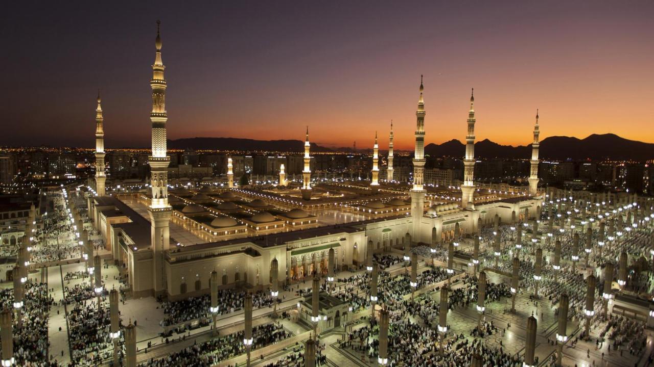 Intercontinental Dar Al Iman Madinah By Ihg Hotel Medina Eksteriør billede The photo depicts an aerial view of a large mosque complex during sunset. The mosque features multiple tall minarets, which are intricately designed and illuminated, giving the scene a stunning appearance as the sky transitions from daylight to dusk.