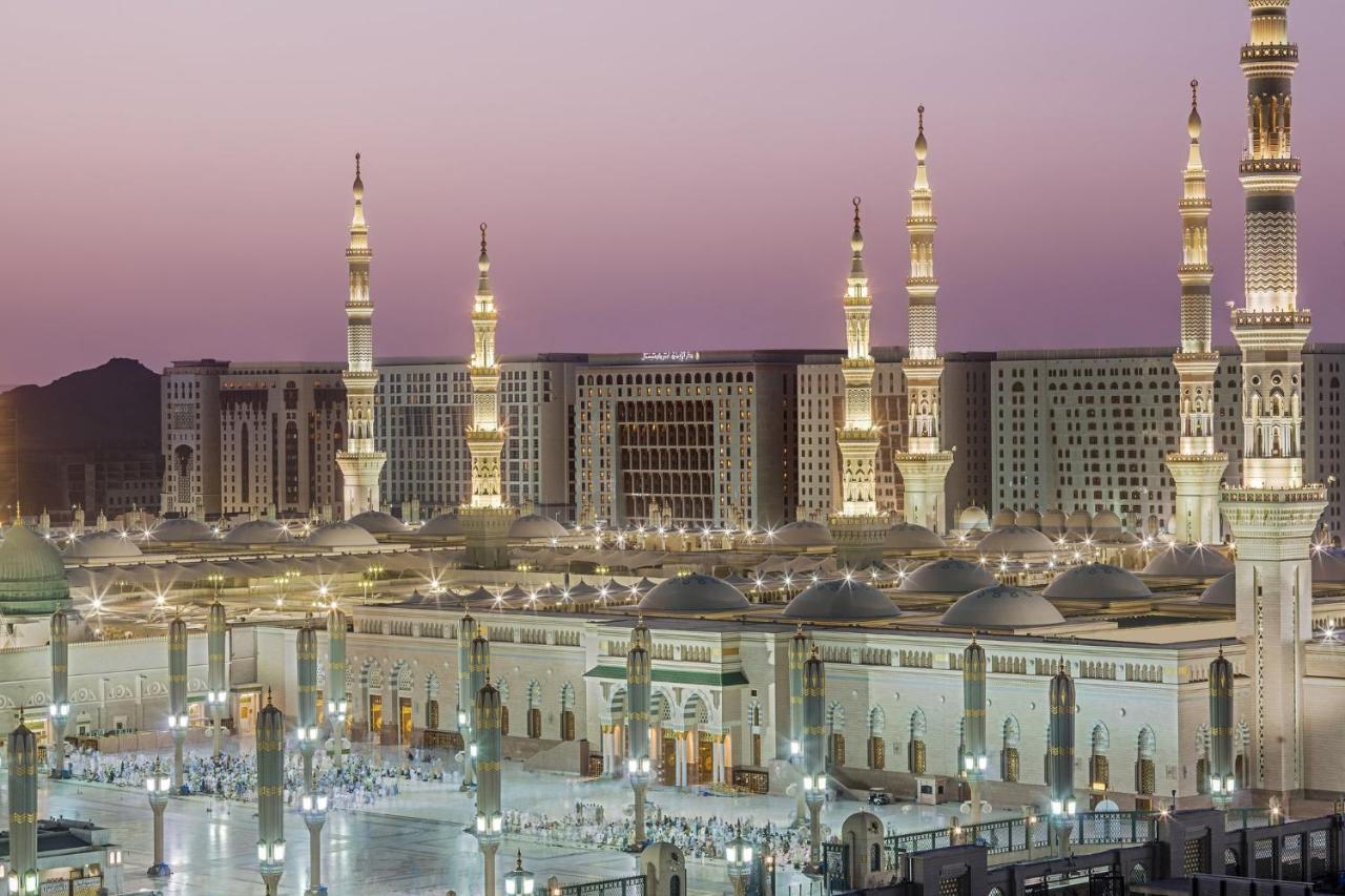 Intercontinental Dar Al Iman Madinah By Ihg Hotel Medina Eksteriør billede The photo shows a view of an expansive mosque complex, featuring several tall minarets that are illuminated against a twilight sky. The structure appears grand and is likely adorned with intricate architectural details. There are people gathered in t