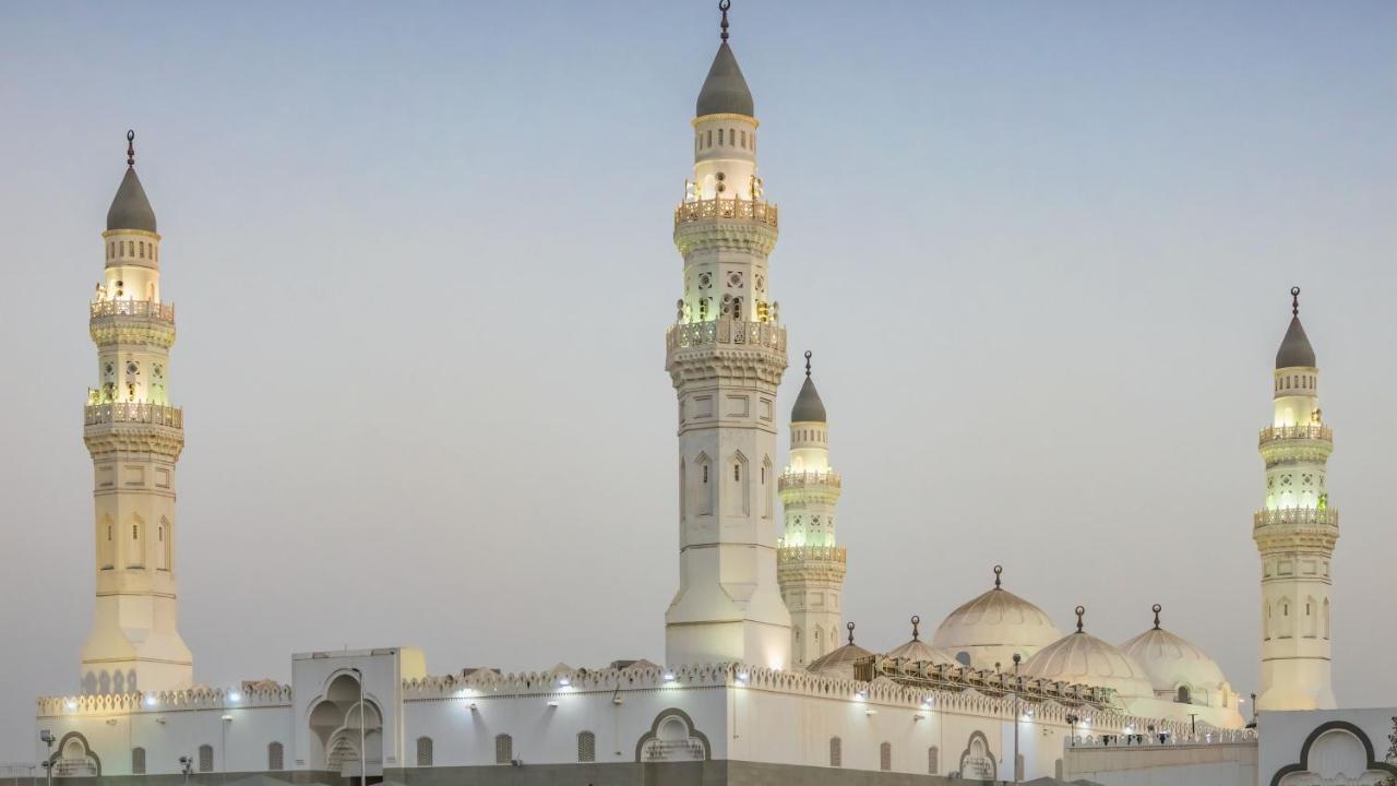 Intercontinental Dar Al Iman Madinah By Ihg Hotel Medina Eksteriør billede The image shows a mosque with distinct architectural features, including tall minarets and domes. The minarets are highlighted, likely lit up in the evening, showcasing intricate design elements typical of Islamic architecture. The overall scene conv