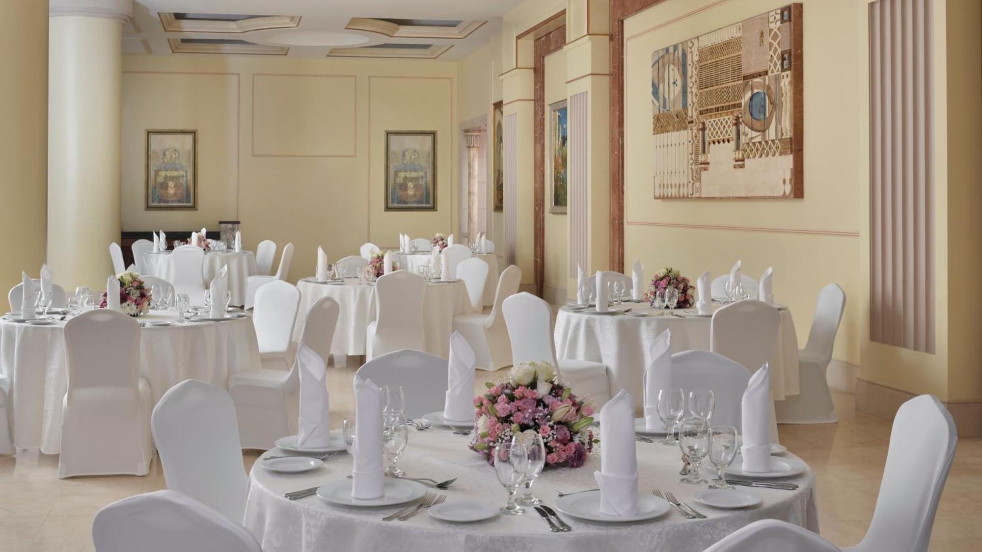 Intercontinental Dar Al Iman Madinah By Ihg Hotel Medina Eksteriør billede The photo shows an elegantly arranged dining area, likely set up for a formal event or reception. There are several round tables covered with white tablecloths, each adorned with floral centerpieces featuring pink flowers. The tables are set with pla