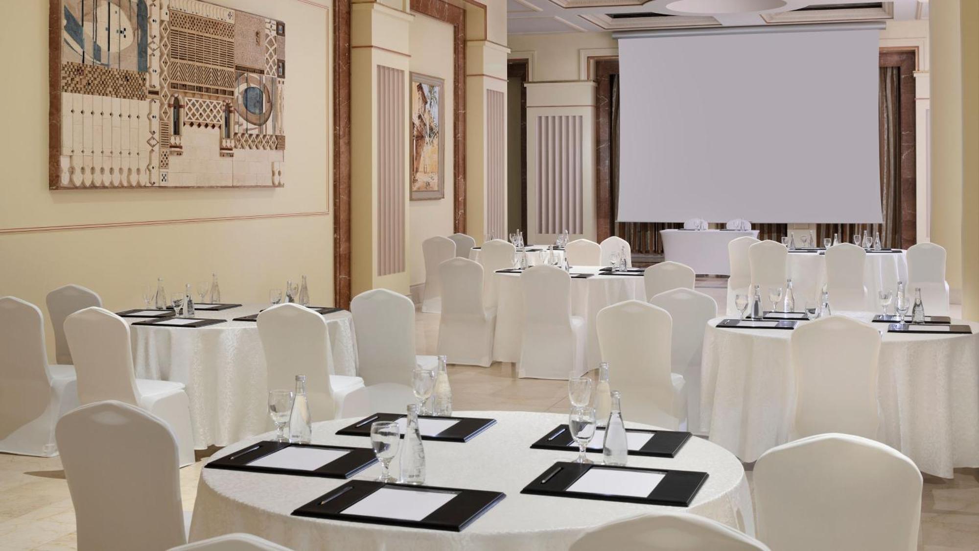 Intercontinental Dar Al Iman Madinah By Ihg Hotel Medina Eksteriør billede The photo depicts an elegantly arranged event space. There are several round tables covered with white tablecloths and surrounded by white chairs. Each table is set with a black folder or menu and a glass of water. The walls are adorned with artworks