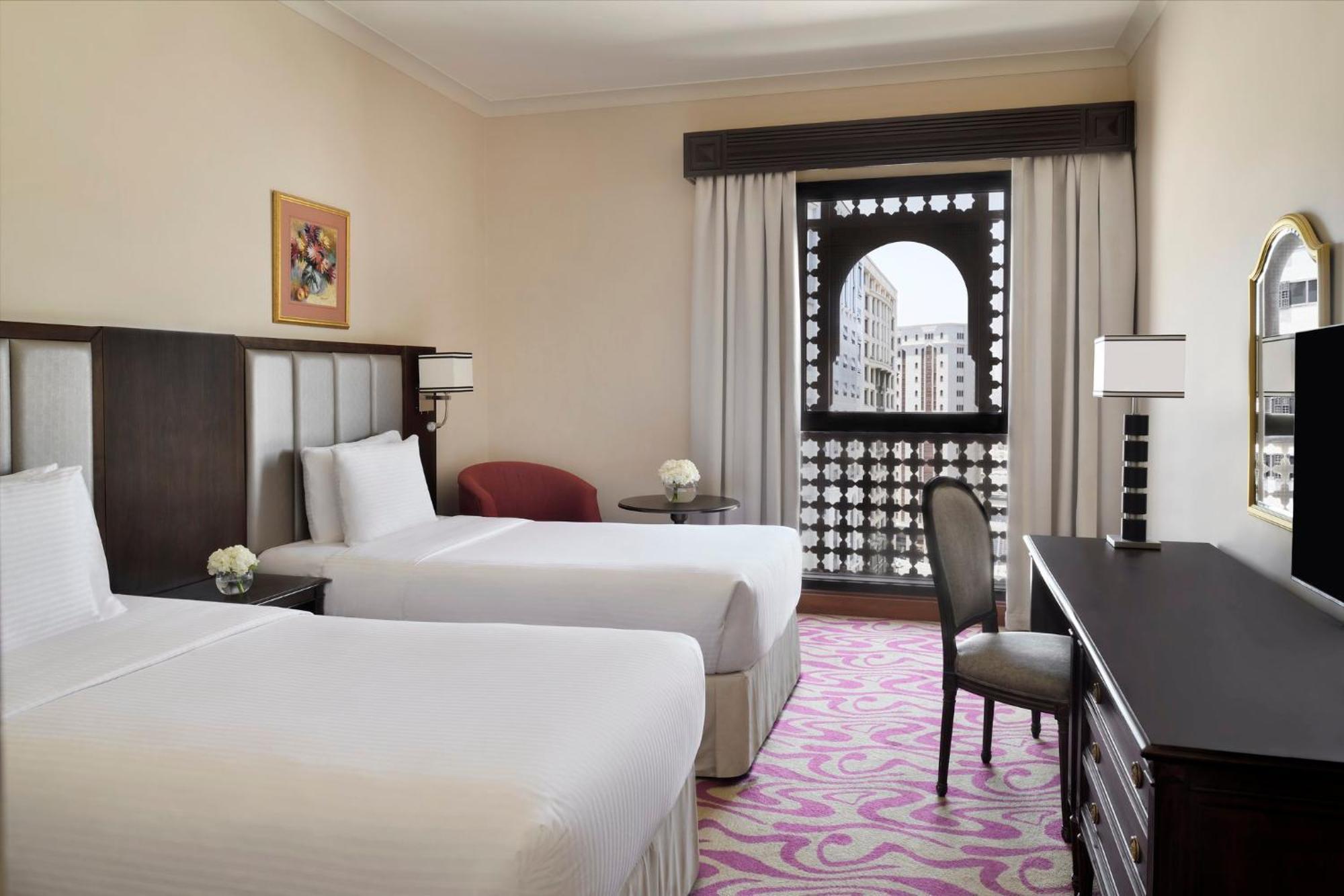 Intercontinental Dar Al Iman Madinah By Ihg Hotel Medina Eksteriør billede The photo shows a hotel room featuring two single beds with white bedding. The room has a light, neutral color scheme and is decorated with minimal furnishings. There's a small seating area with a red chair and a round table, as well as a desk with a