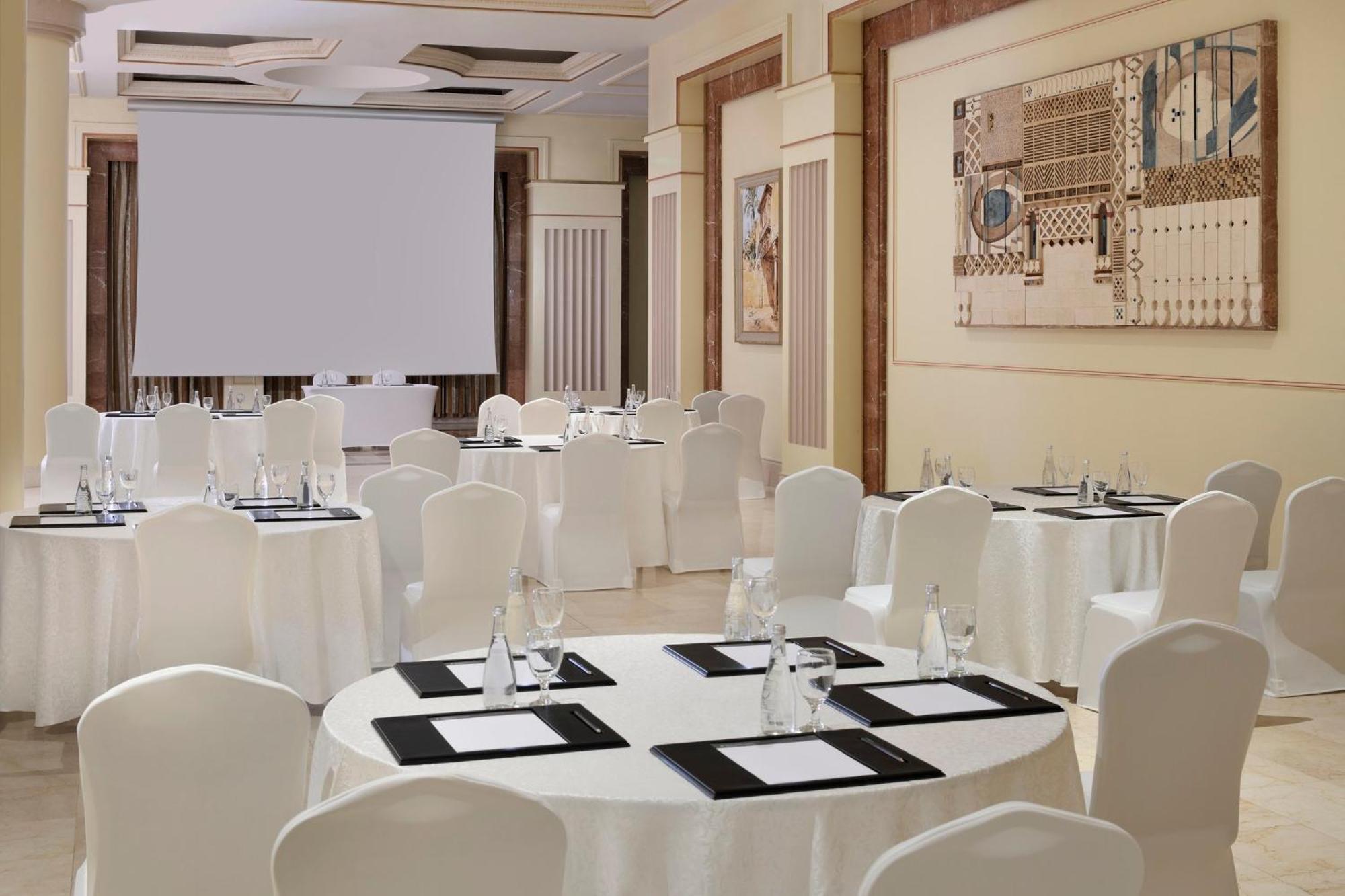 Intercontinental Dar Al Iman Madinah By Ihg Hotel Medina Eksteriør billede The photo shows a well-decorated event space or meeting room. It features several round tables covered with white tablecloths and surrounded by white chairs. Each table has black placemats and glass water bottles, suggesting a formal setup for a gath