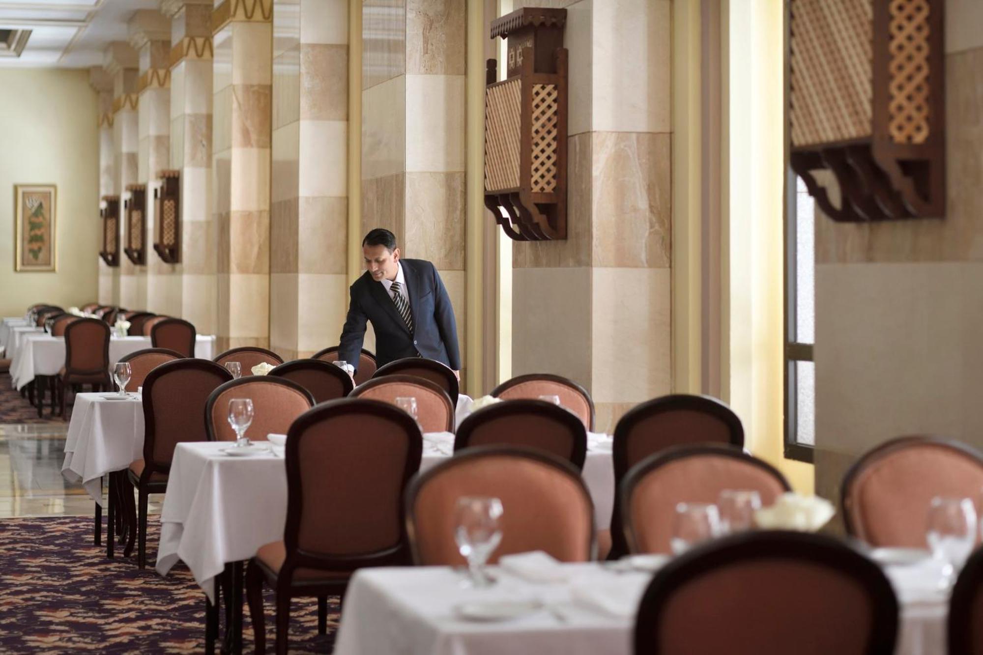 Intercontinental Dar Al Iman Madinah By Ihg Hotel Medina Eksteriør billede The photo depicts a dining area in an elegant restaurant. There are several tables set with white tablecloths, each accompanied by chairs. A waiter, dressed in a formal suit, is seen arranging or tending to one of the tables. The restaurant features 