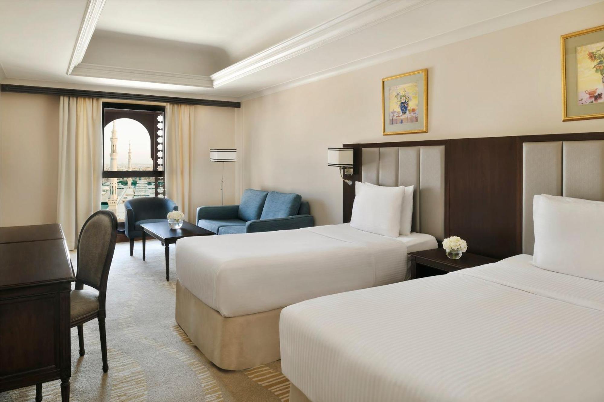 Intercontinental Dar Al Iman Madinah By Ihg Hotel Medina Eksteriør billede The photo shows a well-appointed hotel room featuring two twin beds with white bedding. There is a small desk with a chair, a comfortable blue sofa, and a coffee table in the sitting area. The room has a warm, neutral color scheme, accented by two pa
