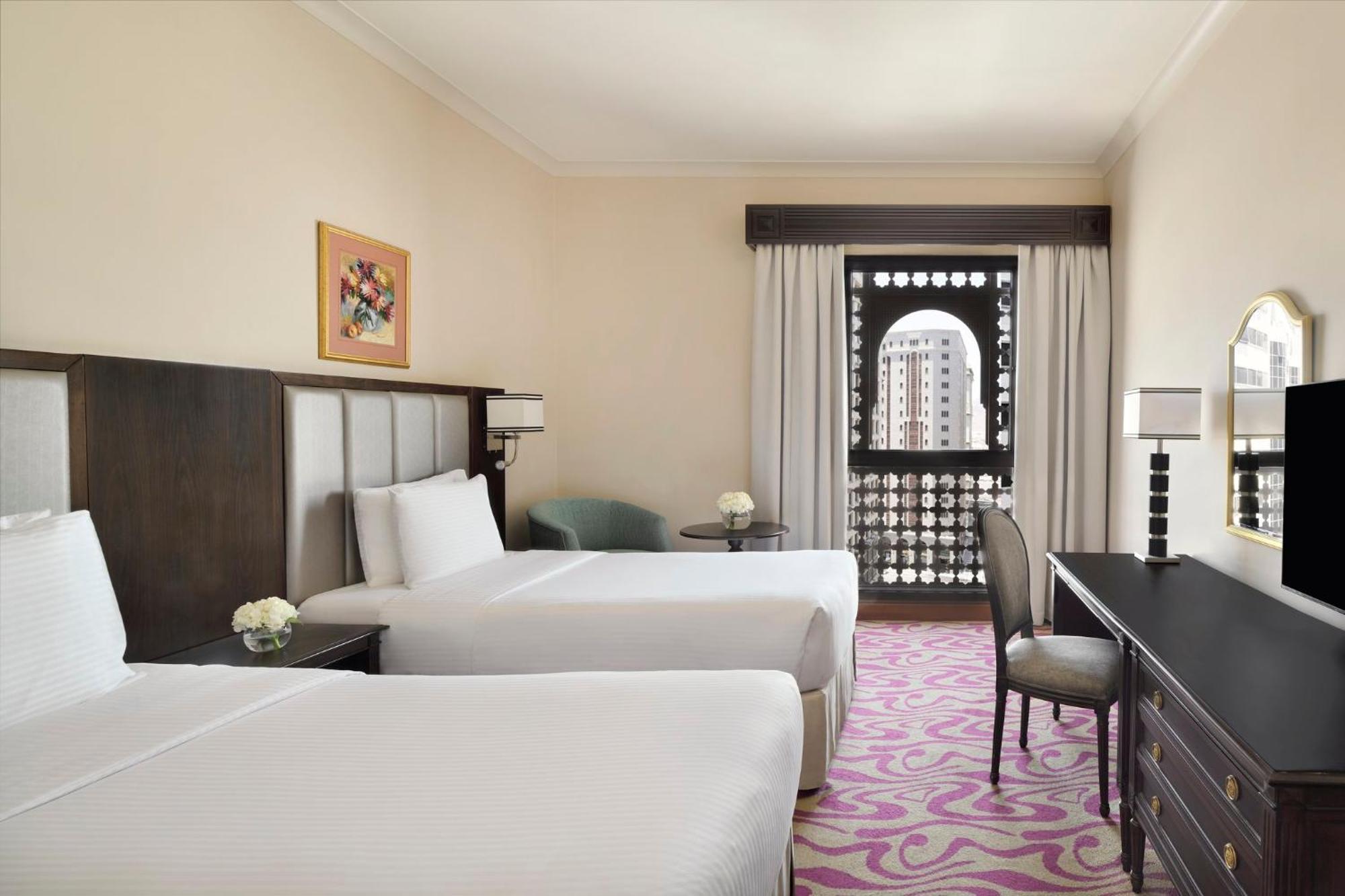 Intercontinental Dar Al Iman Madinah By Ihg Hotel Medina Eksteriør billede The photo shows a hotel room featuring two twin beds with white bedding. There is a decorative piece of art on the wall above one of the beds. A small round table with a flower arrangement is placed next to the beds. A desk and chair are situated aga