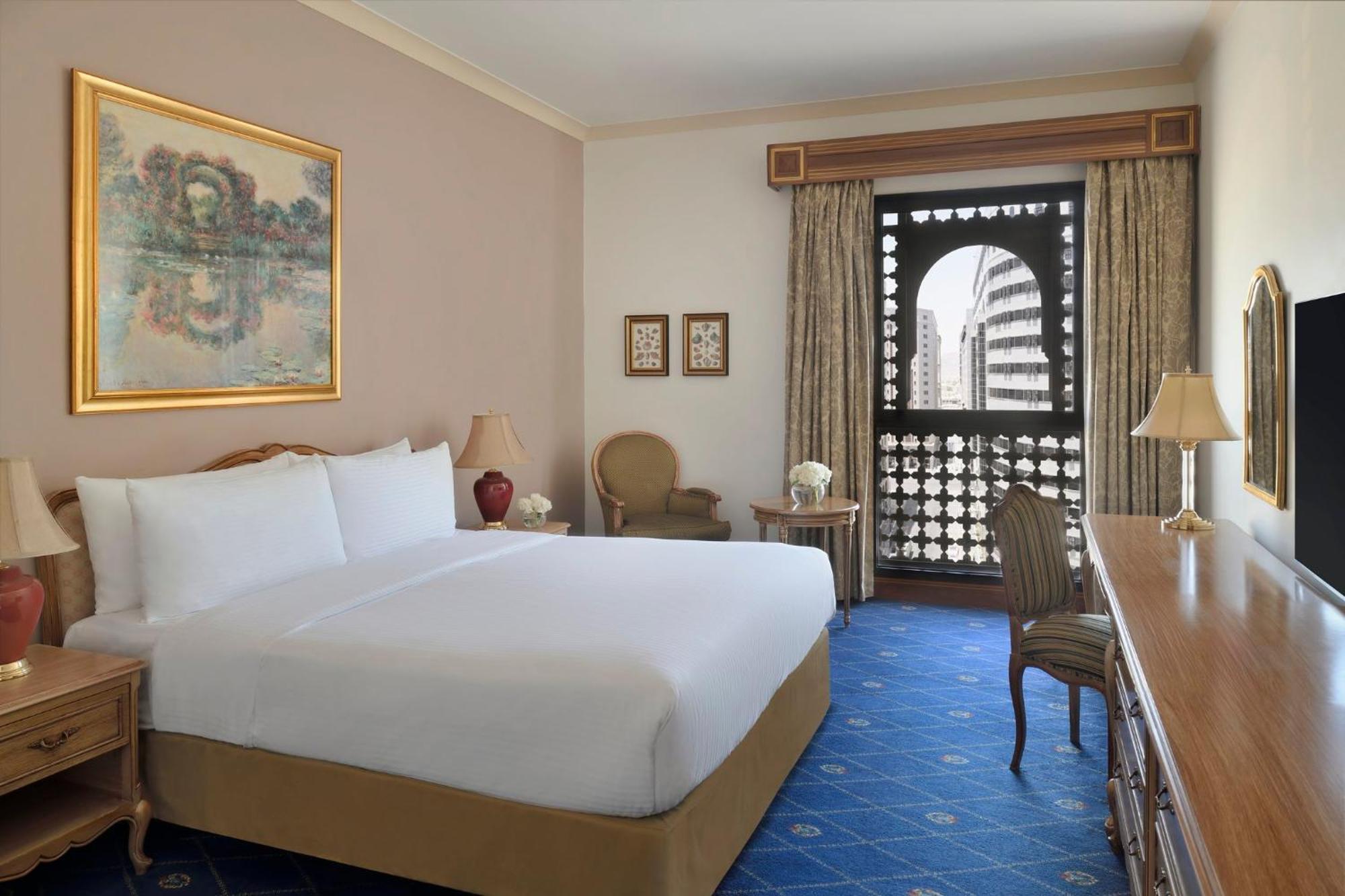 Intercontinental Dar Al Iman Madinah By Ihg Hotel Medina Eksteriør billede The photo shows a hotel room that features a large, neatly made bed with white linens. There is a framed painting on the wall above the bed. To the side, a small seating area includes a chair and a side table. Opposite the bed, there is a wooden desk