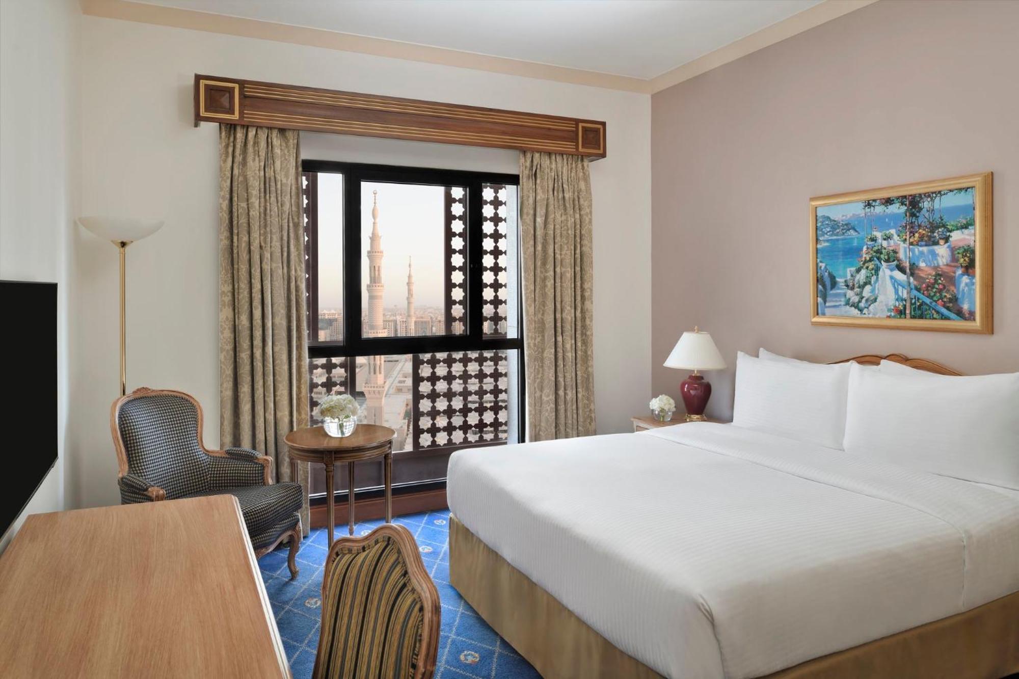 Intercontinental Dar Al Iman Madinah By Ihg Hotel Medina Eksteriør billede The photo shows a hotel room with a modern design. There is a large bed with white linens placed against the wall. Next to the bed, there is a small bedside table with a lamp. A comfortable armchair is situated nearby, accompanied by a wooden desk. T