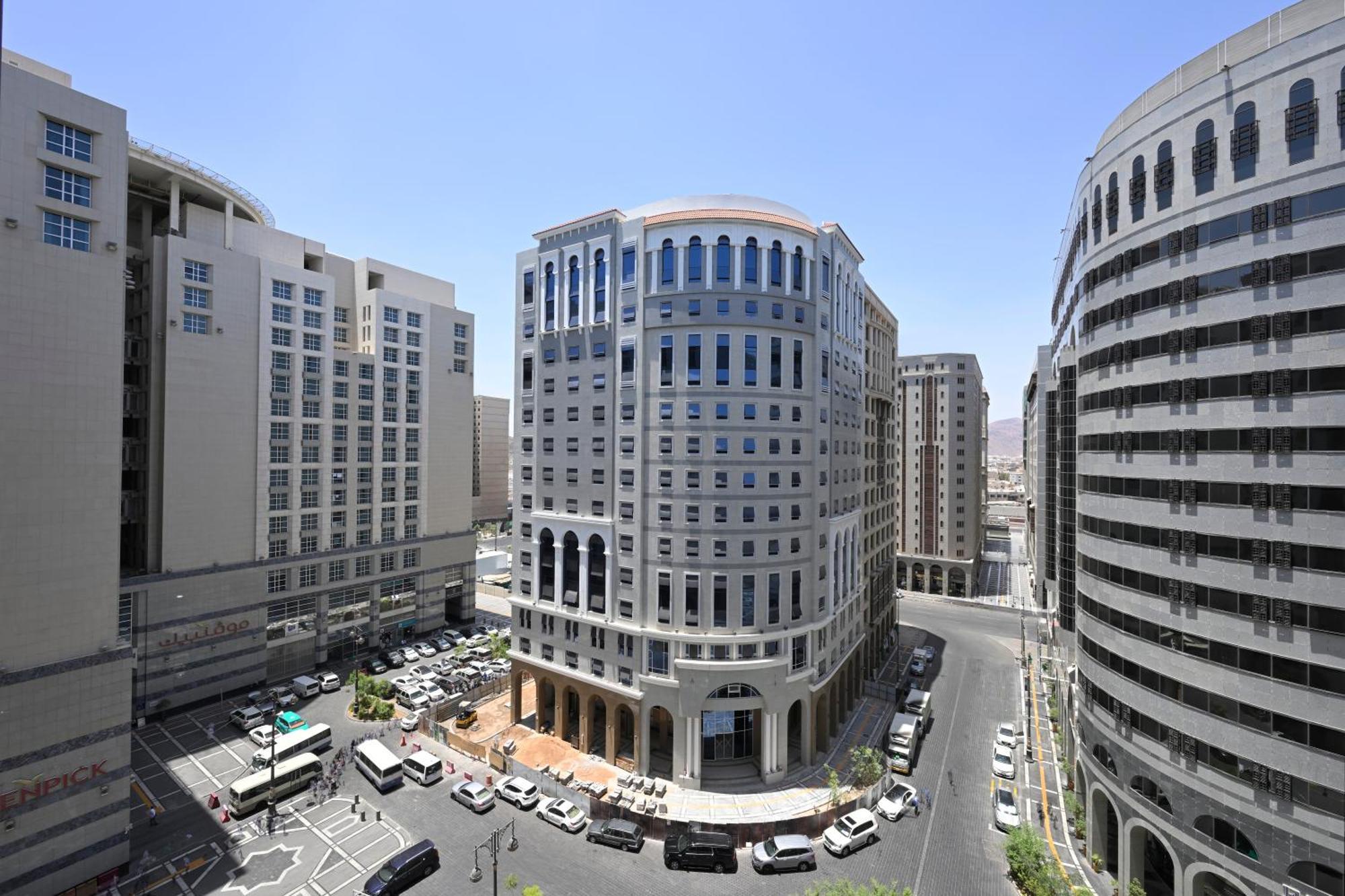 Intercontinental Dar Al Iman Madinah By Ihg Hotel Medina Eksteriør billede The photo shows a modern urban scene featuring several buildings, including a prominent rounded building with a glass façade. This building appears to be situated at an intersection, surrounded by other high-rise structures that have distinct archite