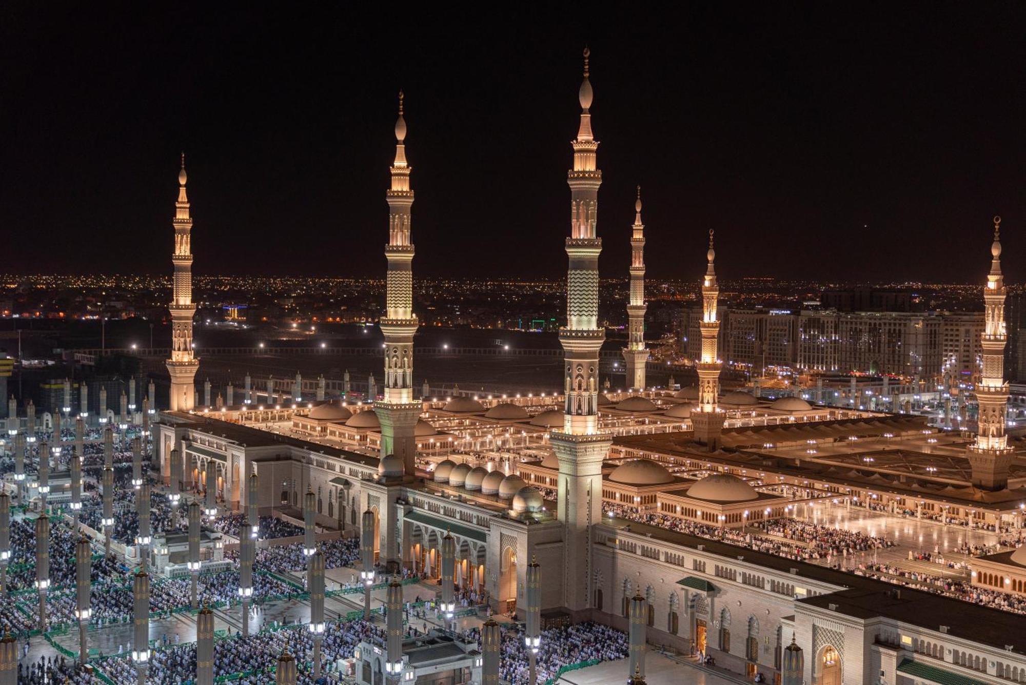 Intercontinental Dar Al Iman Madinah By Ihg Hotel Medina Eksteriør billede The photo depicts a stunning nighttime view of a large mosque complex, prominently featuring tall minarets that are illuminated against the dark sky. The surrounding area is bustling with people, likely worshippers, gathered in the courtyard of the m