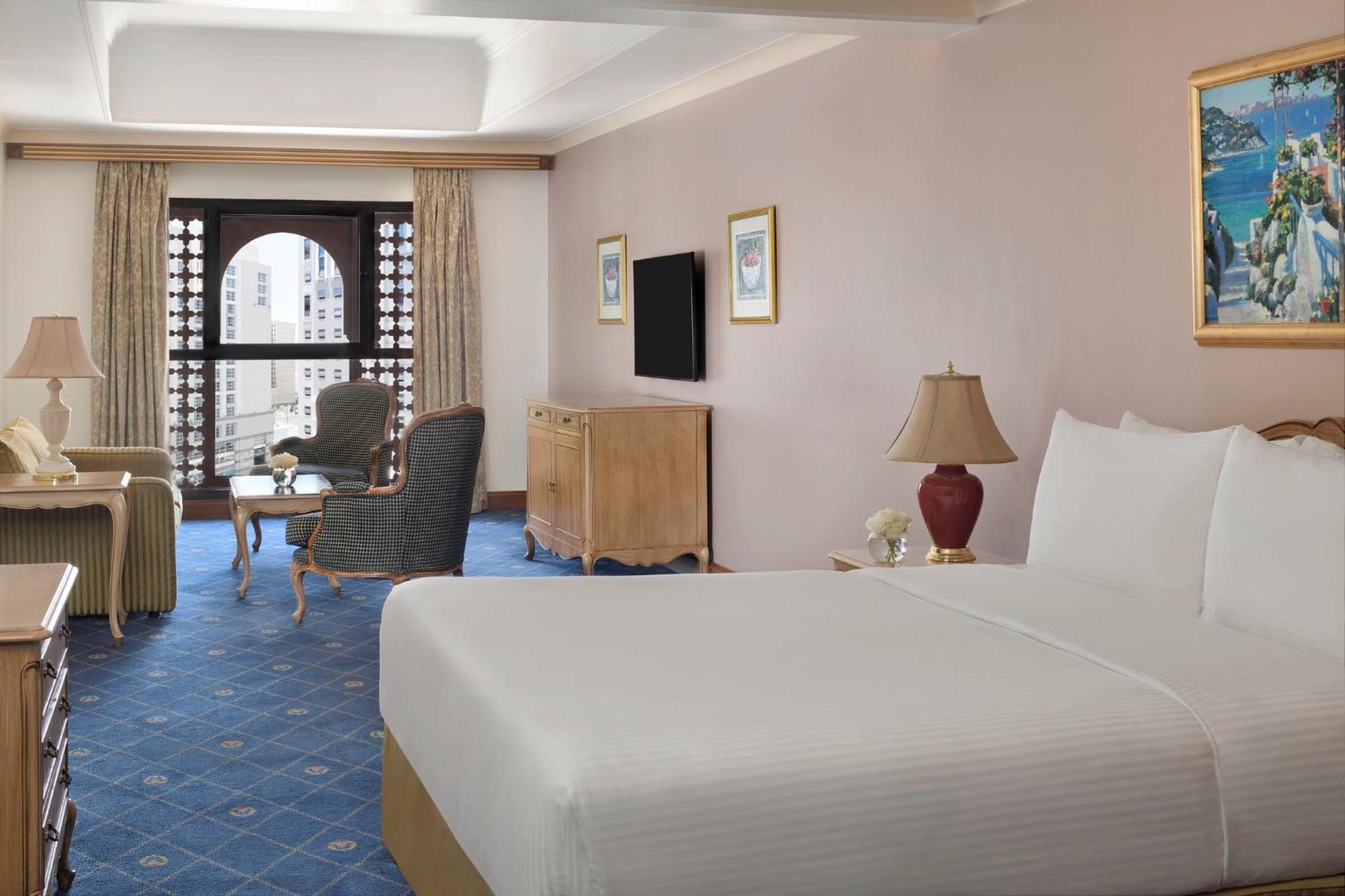 Intercontinental Dar Al Iman Madinah By Ihg Hotel Medina Eksteriør billede The photo shows a well-furnished hotel room. It features a large bed with a white duvet, two bedside lamps, and decorative paintings on the walls. There is a comfortable seating area with two chairs and a small table. A wooden dresser is positioned a