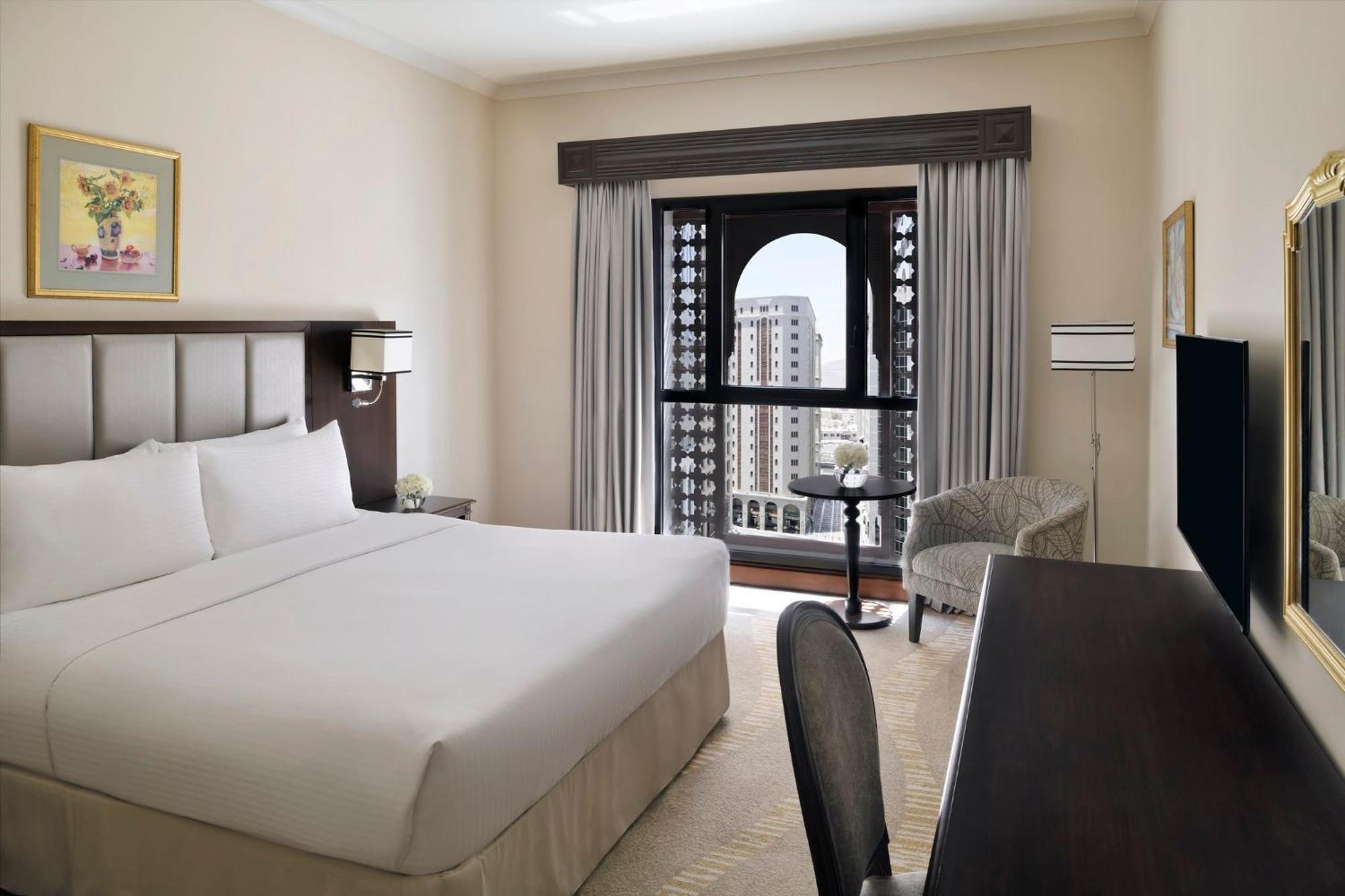 Intercontinental Dar Al Iman Madinah By Ihg Hotel Medina Eksteriør billede The photo shows a hotel room with a modern and minimalistic design. It features a large bed with white linens, a side table, and a comfortable chair near a window. The window has a view of a cityscape, and elegant curtains frame the window. There is 