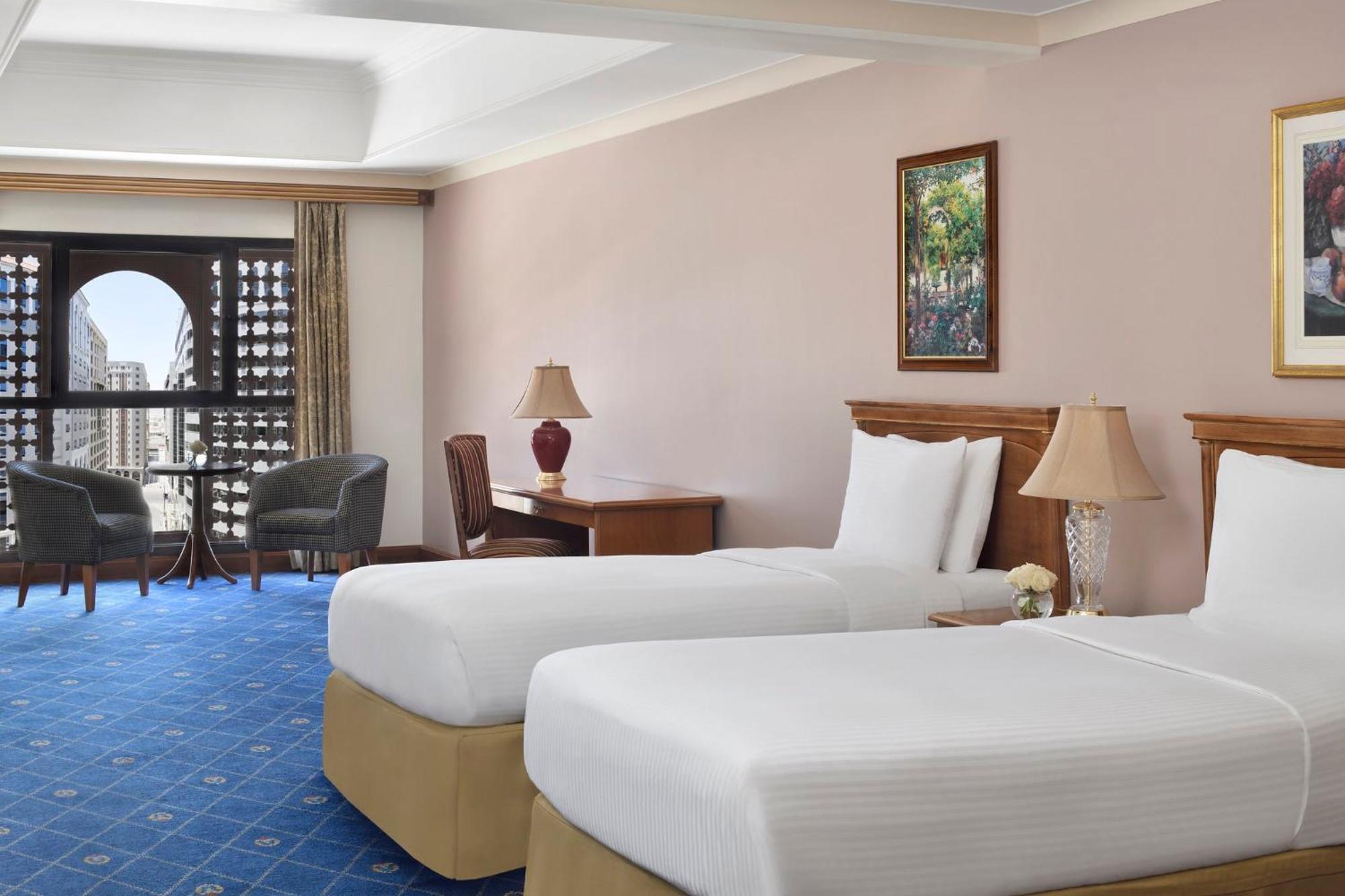 Intercontinental Dar Al Iman Madinah By Ihg Hotel Medina Eksteriør billede The photo shows a nicely designed hotel room featuring two twin beds with white linens and beige bases. In the background, there's a wooden desk and a lamp, along with a pair of comfortable armchairs positioned near a large window. The window showcas