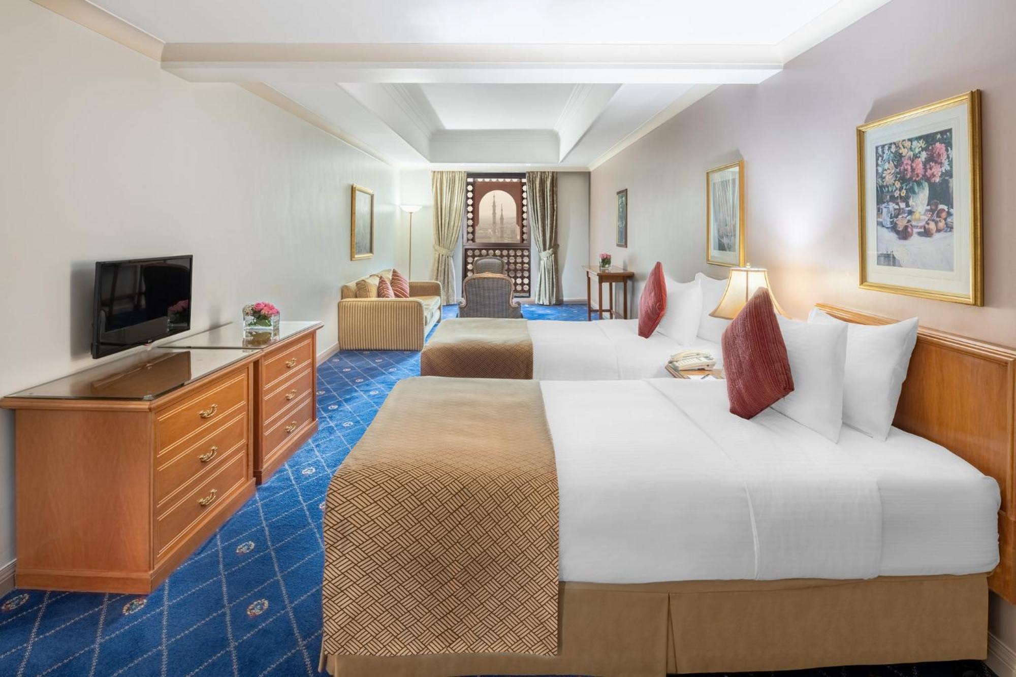 Intercontinental Dar Al Iman Madinah By Ihg Hotel Medina Eksteriør billede The photo shows a hotel room featuring two beds with white linens and decorative pillows. There is a wooden dresser with a television on it. The flooring has a blue carpet with a pattern, and the walls are painted in a light color. The room is well-l