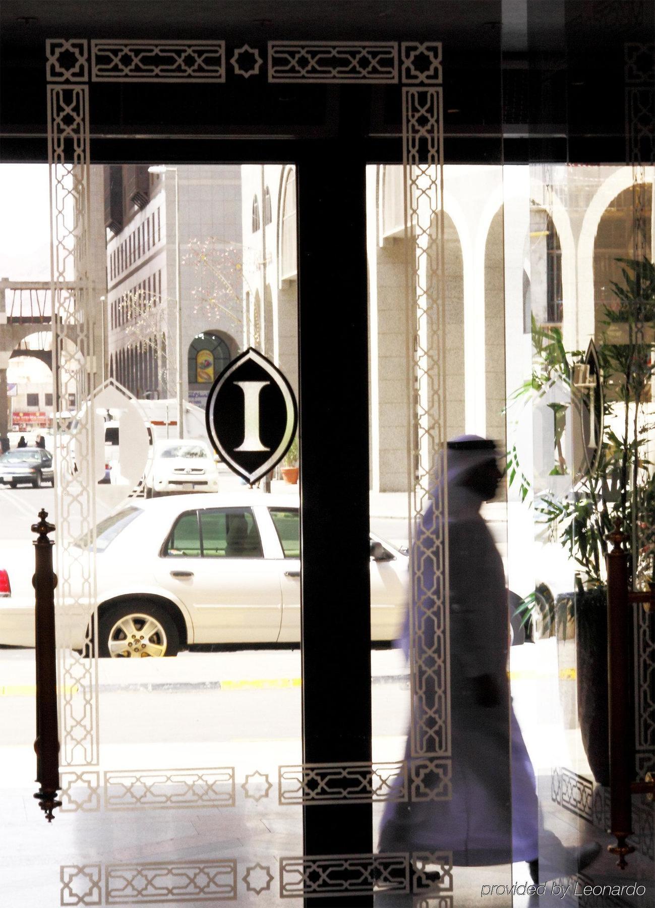 Intercontinental Dar Al Iman Madinah By Ihg Hotel Medina Interiør billede The photo shows a view through a glass door or window, featuring an elegant design with a prominent "I" symbol displayed. Through the glass, there are some parked vehicles visible, including a white car. In the foreground, a shadowy figure of a perso