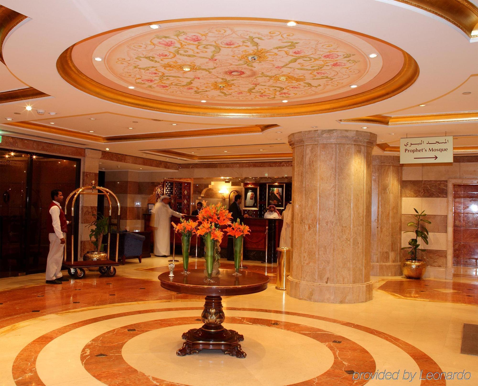 Intercontinental Dar Al Iman Madinah By Ihg Hotel Medina Interiør billede The photo depicts a luxurious hotel lobby. The ceiling features an ornate design with floral patterns, adding elegance to the space. In the center of the lobby, there is a decorative table with a bowl or vase of bright orange flowers. The flooring is