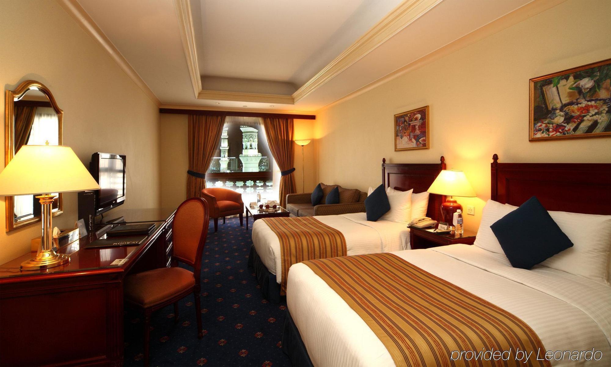 Intercontinental Dar Al Iman Madinah By Ihg Hotel Medina Værelse billede The photo shows a hotel room featuring two beds with white linens and brown striped throws. There is a wooden desk and chair, alongside a television. The walls are painted in a warm tone, and there are decorative paintings. Large windows with curtain