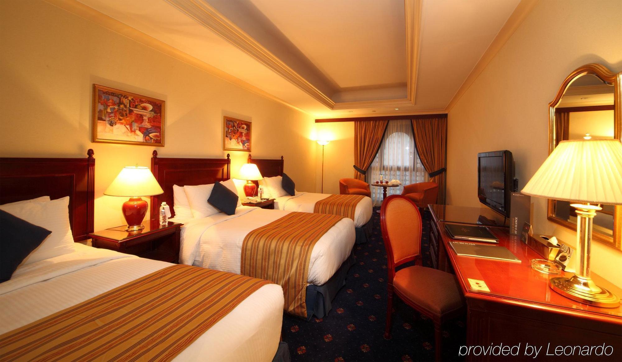 Intercontinental Dar Al Iman Madinah By Ihg Hotel Medina Værelse billede The photo shows a hotel room with a warm and inviting atmosphere. There are two beds with white and gold bedding, complemented by decorative pillows. Each bed has a bedside lamp. In the background, there's a small seating area with a couple of chairs