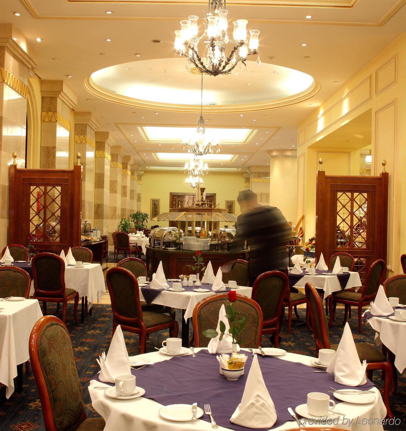 Intercontinental Dar Al Iman Madinah By Ihg Hotel Medina Restaurant billede The photo depicts an elegant restaurant interior. It features round tables covered with white tablecloths, each set with plates, cutlery, and folded napkins arranged into triangles. The space has a warm, inviting atmosphere with a mix of decorative e