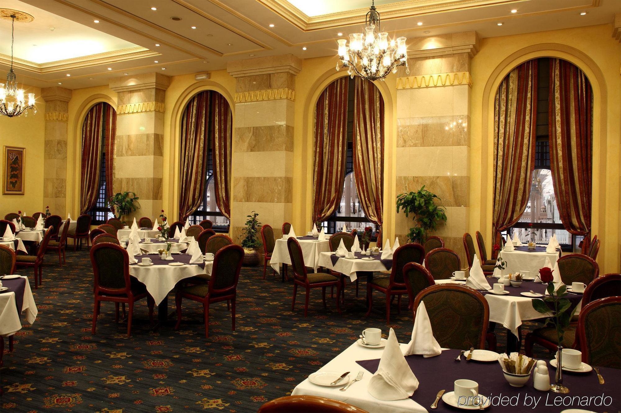 Intercontinental Dar Al Iman Madinah By Ihg Hotel Medina Restaurant billede The photo depicts an elegant restaurant interior. The space features ornate decor with tall windows draped in rich, patterned curtains. Tables are neatly set with white tablecloths and napkins, and there are plush chairs arranged around them. The flo