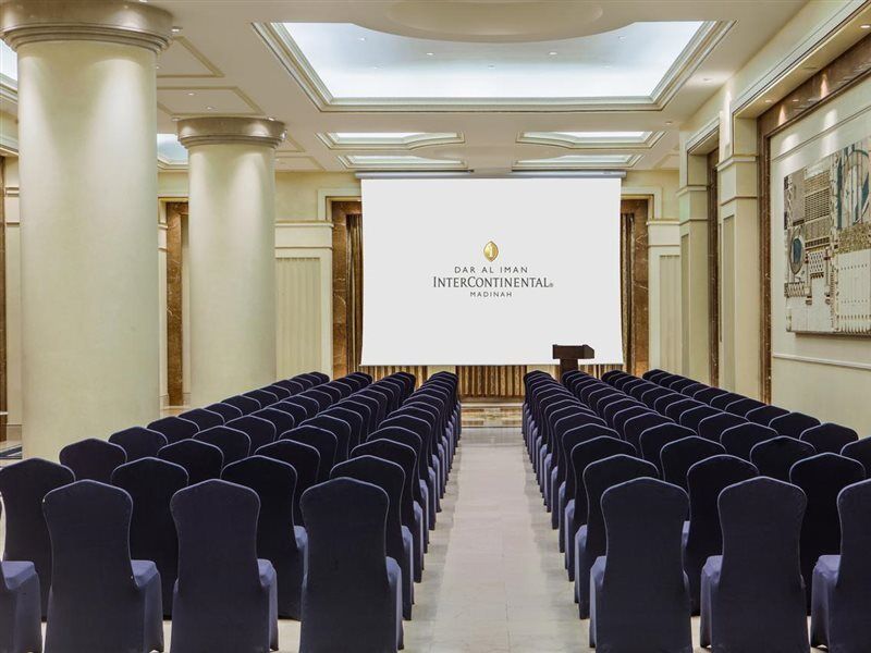 Intercontinental Dar Al Iman Madinah By Ihg Hotel Medina Eksteriør billede The photo shows a conference or meeting room set up. There are rows of dark blue chairs arranged facing a large screen at the front of the room. The screen displays the logo of the Intercontinental hotel, indicating that this space is likely intended