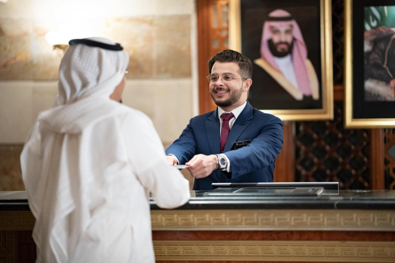 Intercontinental Dar Al Iman Madinah By Ihg Hotel Medina Eksteriør billede The photo shows a hotel reception area. A male receptionist, dressed in a navy suit with a tie, is smiling and handing an item to a guest who is wearing traditional white attire, possibly a kandura or thobe. The background features elegantly decorate