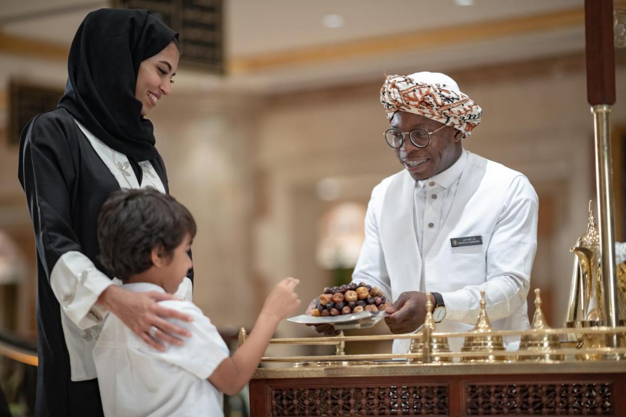 Intercontinental Dar Al Iman Madinah By Ihg Hotel Medina Eksteriør billede The photo depicts a warm interaction between three people. A woman, dressed in a traditional black attire and a headscarf, stands beside a young boy who is reaching out towards a plate of dates held by a smiling man in a white traditional outfit with